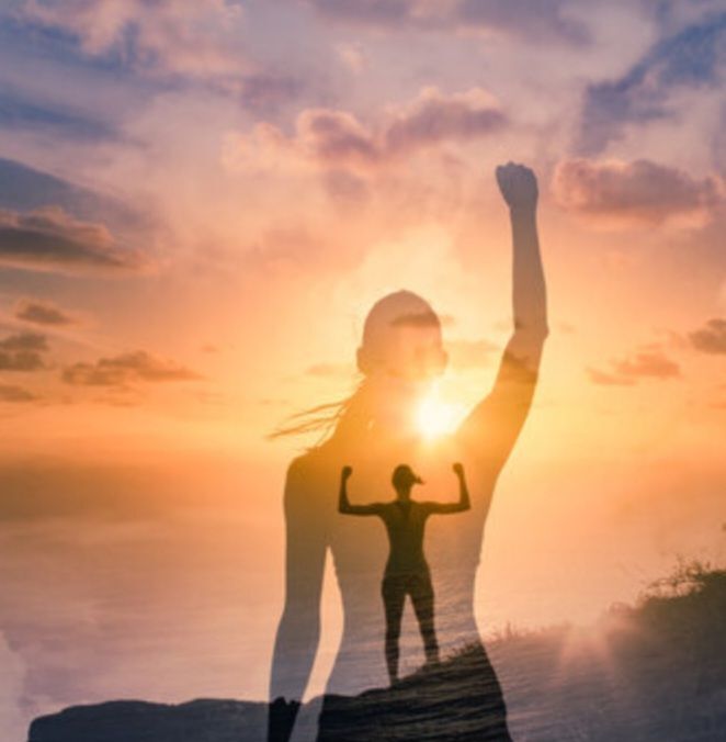 Women standing on a hill overlooking the water with the sun setting and flexing with her shadow is holding one arm up in a fist